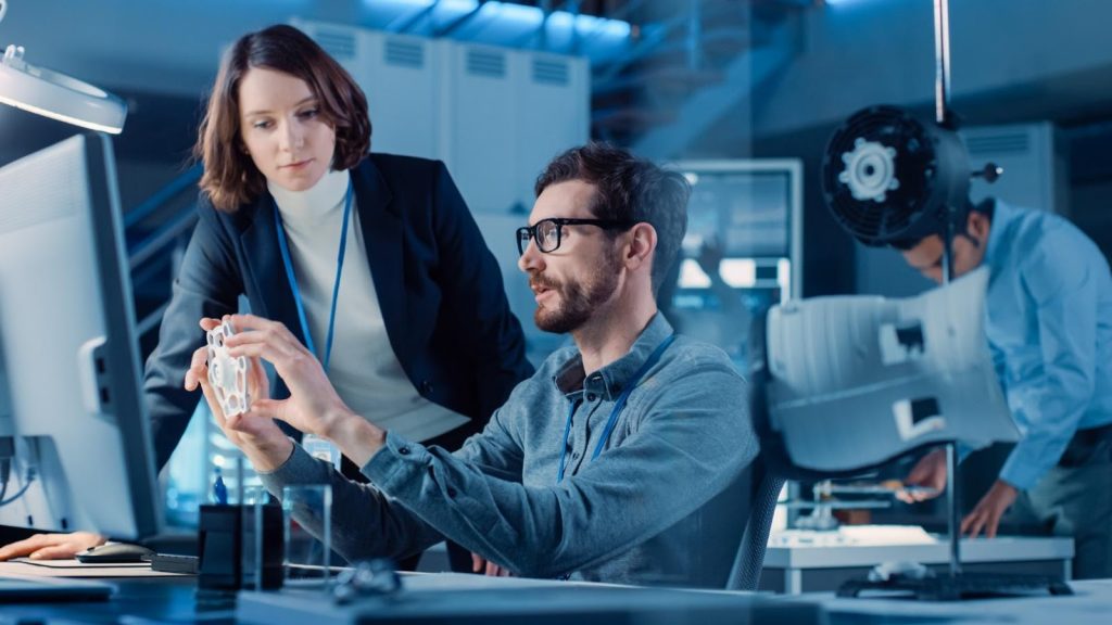 a man peering at a small plastic shape while a coworker watches