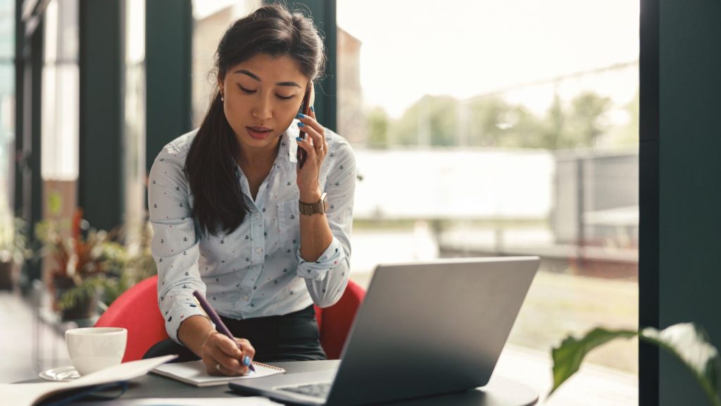 Office worker on the phone and writing notes in front of a laptop