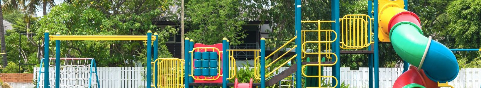 A colorful playground with a slide, swings, and climbing structures on a grassy field. Trees and a white fence are in the background under a blue sky.