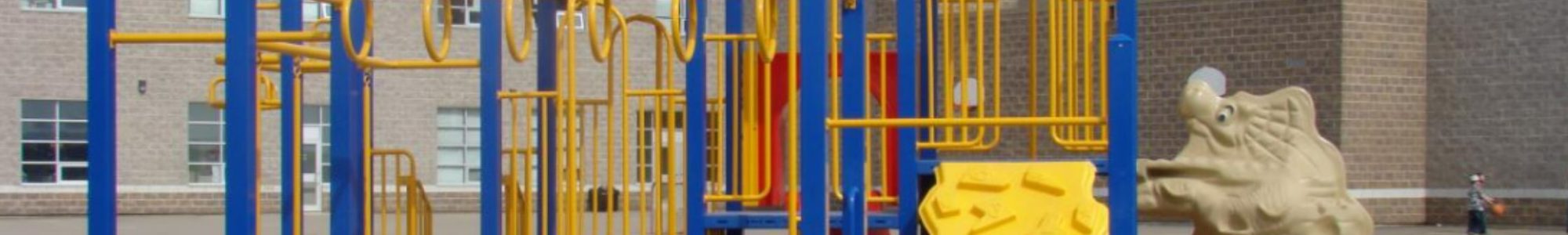 Playground with colorful climbing structures, slides, and ladders in yellow and blue. Background features a brick school building. Sunny day.